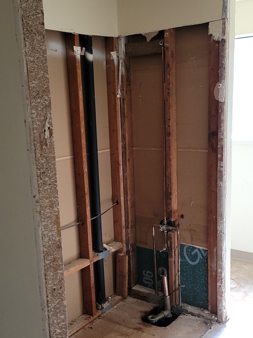 Empty tub alcove stripped to bare studs, with the shower valve and drain pipes exposed and a bright room beyond the doorway