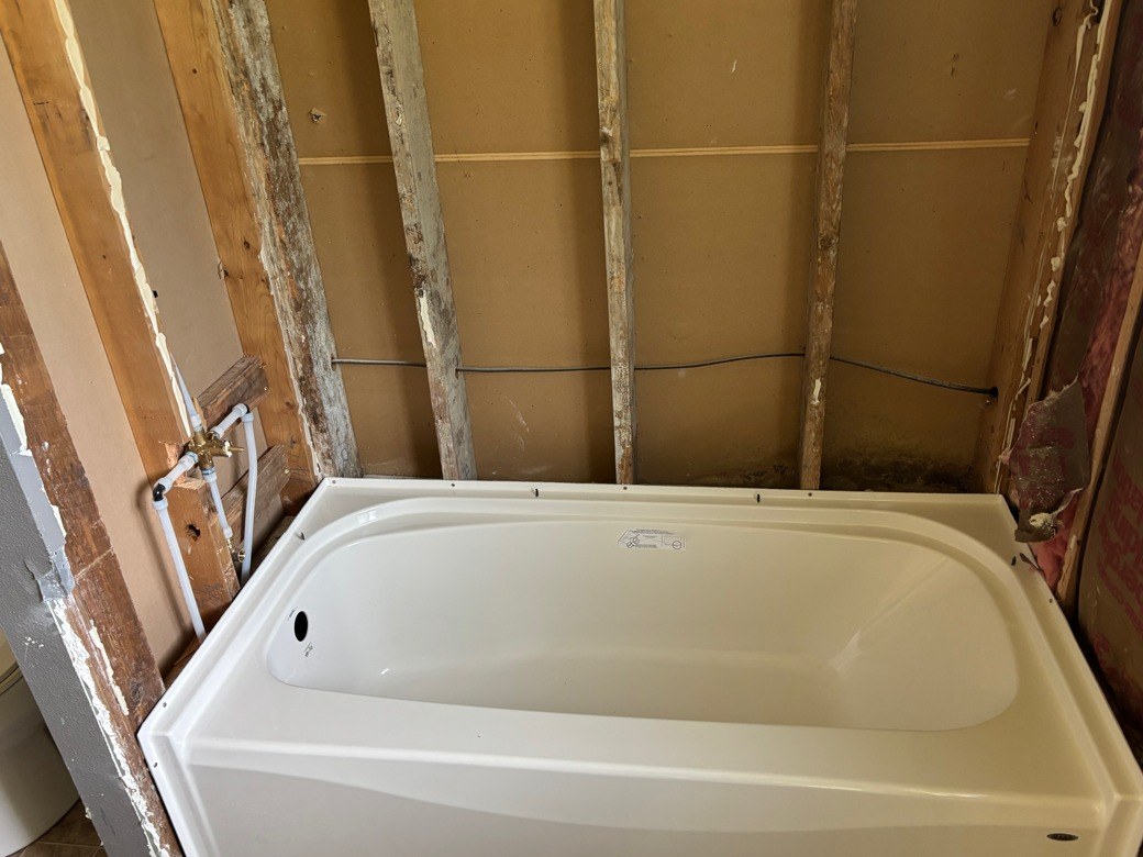 White bathtub set into a stripped bathroom alcove against bare wood studs, with new water lines rising from the wall at its left end