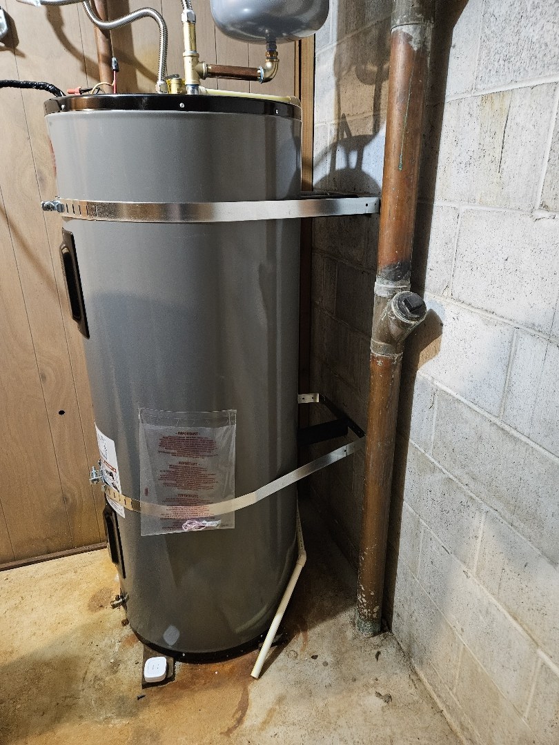 Gray electric water heater held by two metal straps against a cinder-block wall, with copper pipes beside it and a white relief pipe running down toward the floor