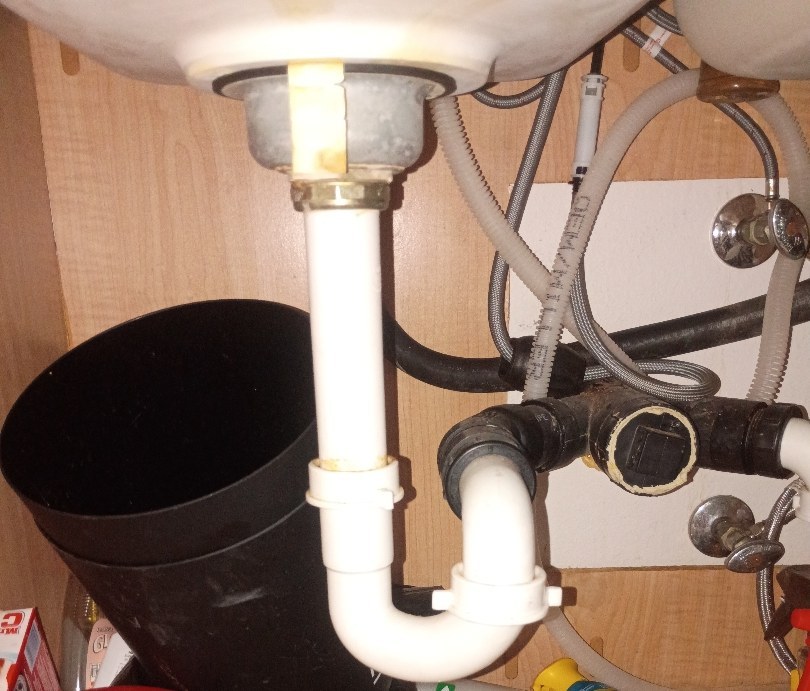 White PVC P-trap and tee under the left basin of a double kitchen sink, beside the black discharge elbow of a garbage disposal