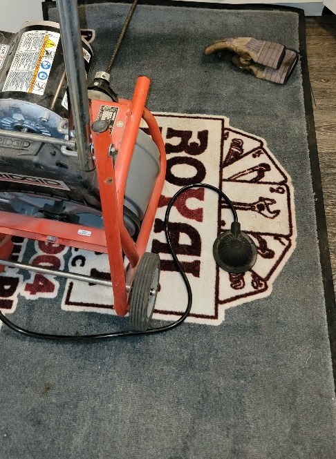 Orange-framed drain cleaning machine standing on a gray floor mat printed with a red and white logo, its cable running up toward the sink
