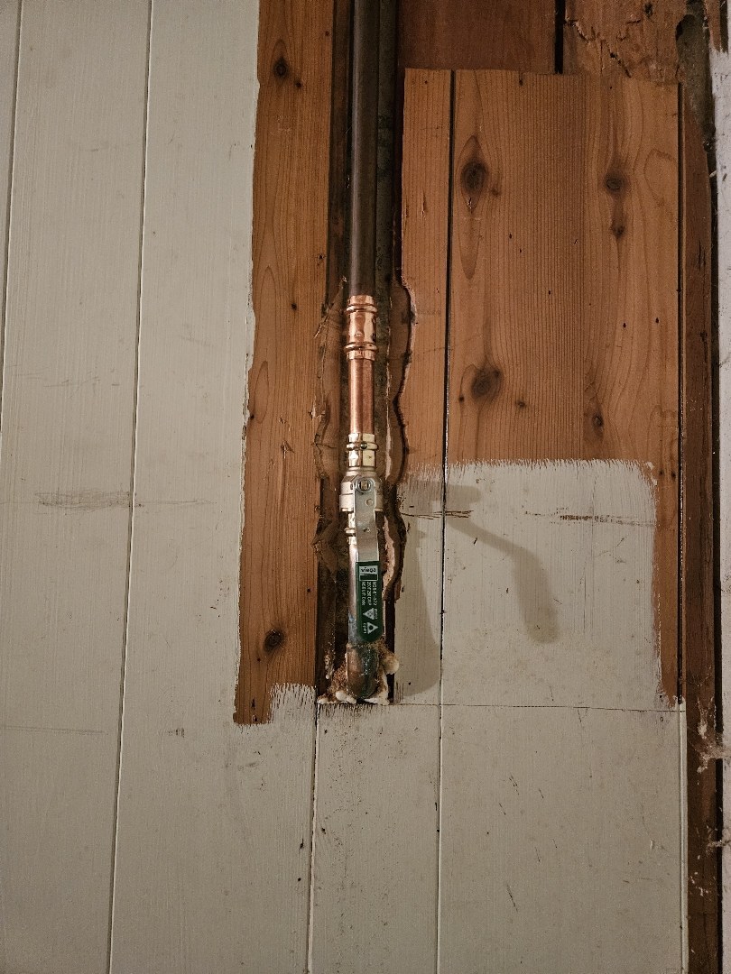 A new valve on a vertical copper water line inside a cut-out in white-painted wood paneling
