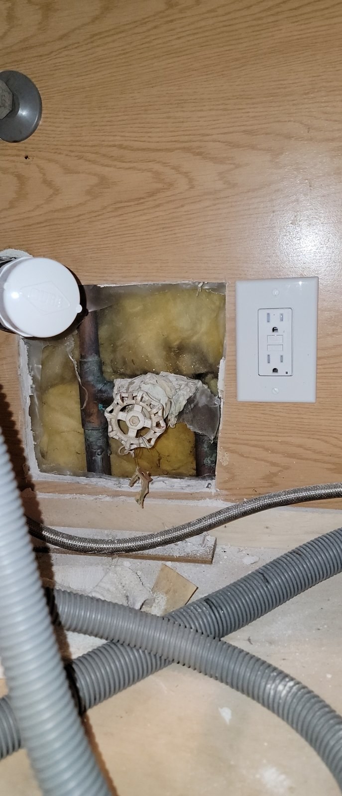 Old round-wheel gate valve on copper pipe inside a cut-out behind a kitchen sink cabinet, next to a wall outlet