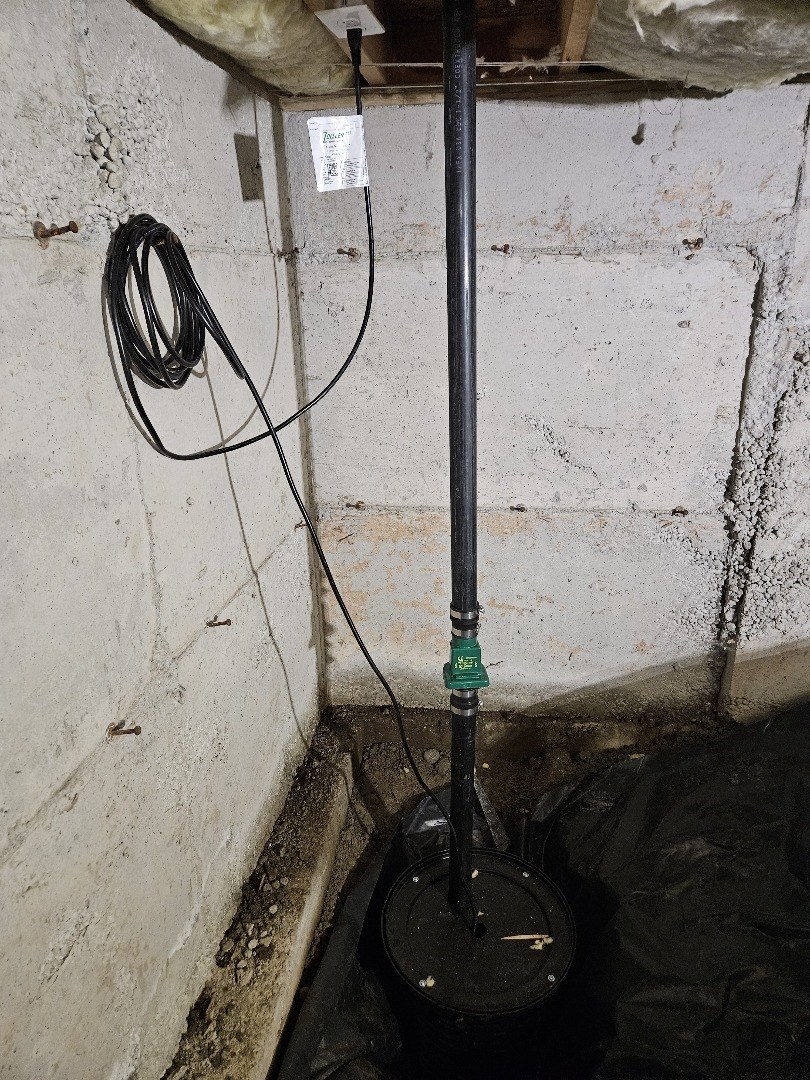 Black sump basin lid set into the floor of a crawl space corner, with a rigid discharge pipe, a green check valve and a coiled power cord against a concrete wall