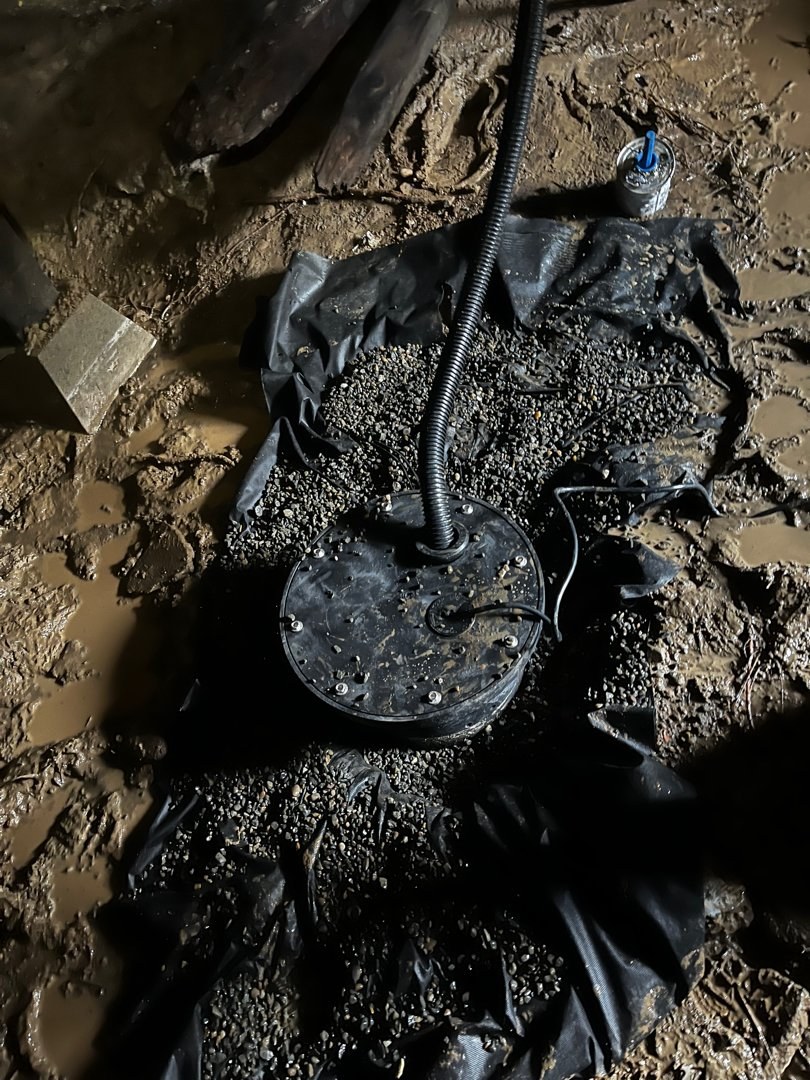 Round black sump basin lid set in pea gravel over black filter fabric on a muddy crawl-space floor, with a corrugated discharge hose rising from it