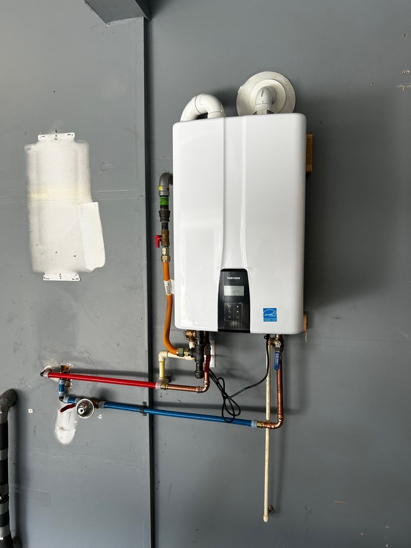White wall-mounted tankless water heater on a gray garage wall, with red and blue water lines, a yellow gas connector and two white vent pipes rising from the top