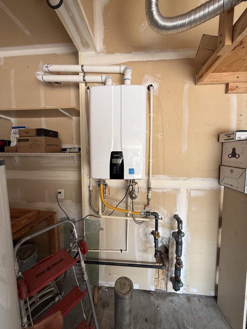 Wall-hung white tankless water heater in a garage with white PVC vent pipes above it and a yellow gas line, water lines and isolation valves below
