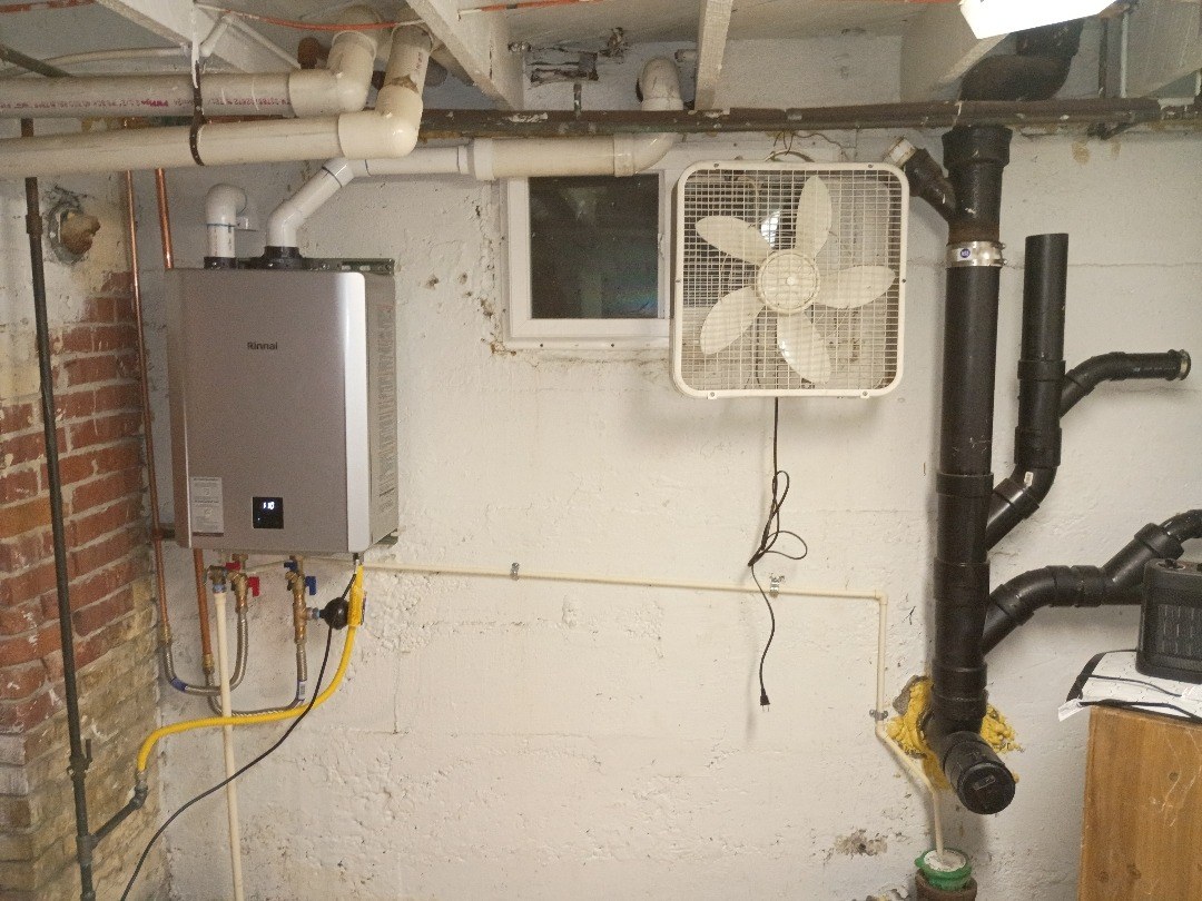 Wall-mounted tankless water heater beside an old brick wall in an unfinished basement, with a yellow flexible gas line below it and white vent pipes along the ceiling